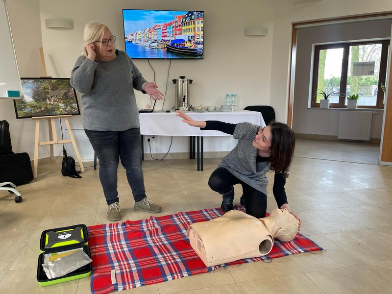 Starsza kobieta stoi rozmawiając przez telefon komórkowy, obok klęczy dziewczyna nad fantomem, opisując sytuacje starszej Pani