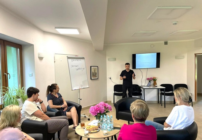 Szkolenie w pomieszczeniu z telewizorem i tablicą. Prelegent stoi przodem do kadru słuchacze siedzą odwróceni do prelegenta