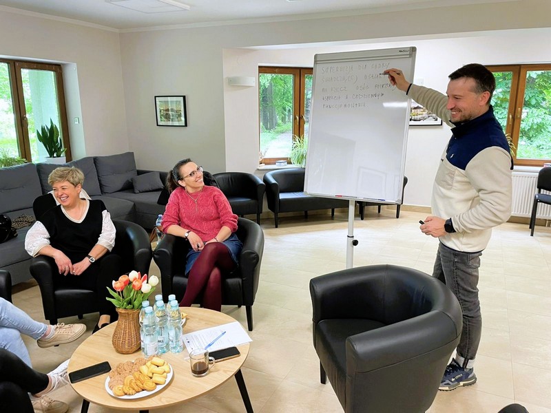 Prelegent pokazujący na tablicę i dwie słuchaczki szeroko uśmiechniete