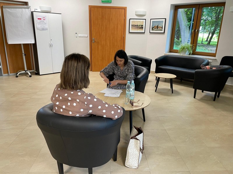 Dwie kobiety siedzą przy małym stoliku na wygodnych skórzanych fotelach. Rozmawiają jedna z n ich wypełnia dokumenty w tle szafa z tablicą oraz drzwi i okno