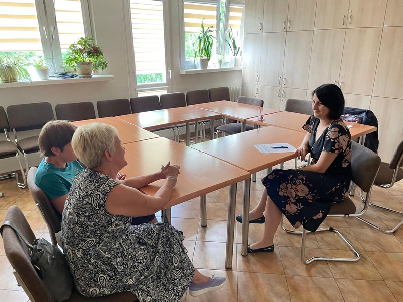 Trzy kobiety siedza w pomieszczeniu przy długim stole ułożonym w prostokąt. Rozmawiają. w tle okna i ściana którą pokrywaja szafki