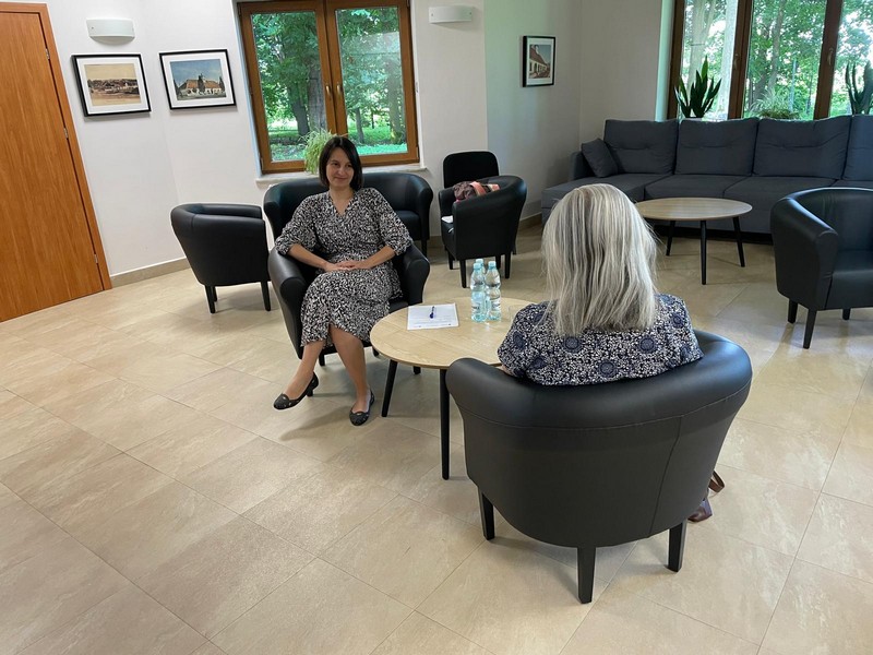 Dwie kobiety siedzą przy stoliku na fotelach i rozmwiają w tle okna z widokiem na zielony park