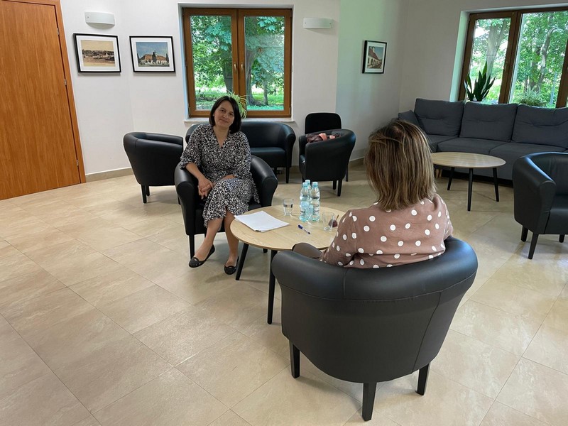 Dwie kobiety siedzą przy małym stoliku na wygodnych skórzanych fotelach. Rozmawiają jedna z n ich wypełnia dokumenty w tle szafa z tablicą oraz drzwi i okno