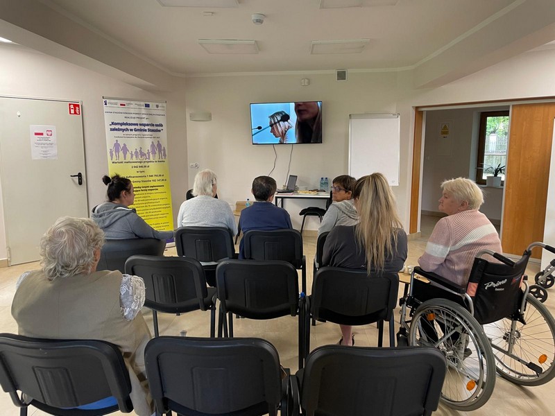 Grupa ludzi siedzących na krzesłach ogląda wspólnie film wyświetlany na telewizorze- jedna z osób siedzi na wózku inwalidzkim