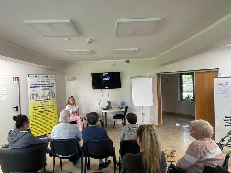 Na zdjęciu grupa ludzi siedzi na krzesłach jedna z osób siedzi na wózku inwalidzkim- Oglądają film wyświetlany na telewizorze-przed nimi stoi Pani w błękitnym swetrze