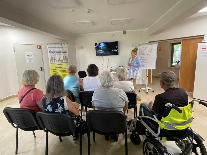 Na zdjęciu grupa ludzi siedzi na krzesłach jedna z osób siedzi na wózku inwalidzkim- Oglądają film wyświetlany na telewizorze-przed nimi stoi Pani w błękitnym swetrze