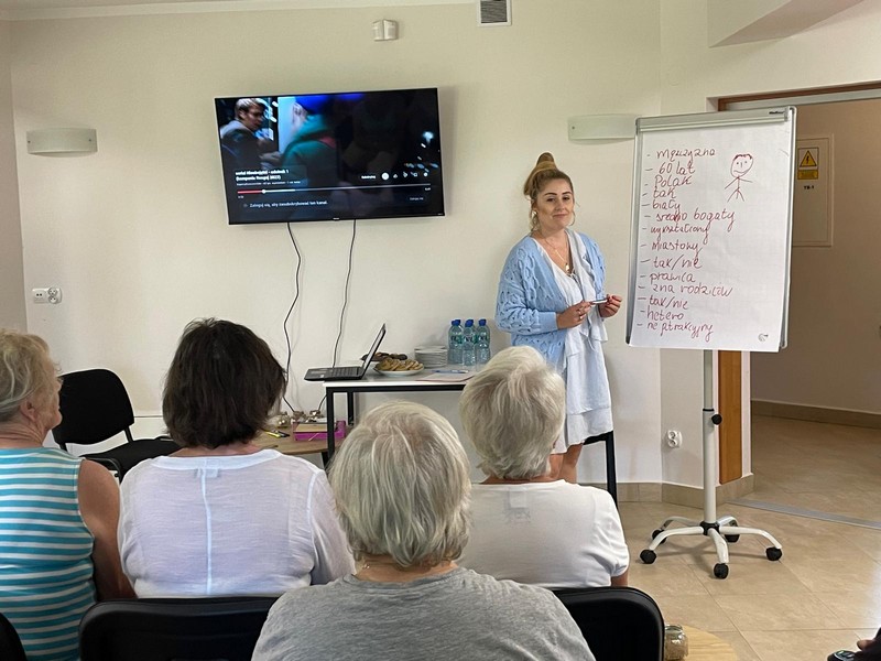 Na zdjęciu stoi Pani w błękitnym swetrze przy tablicy wypisując słowa, przed nią siedzi grupa kobiet