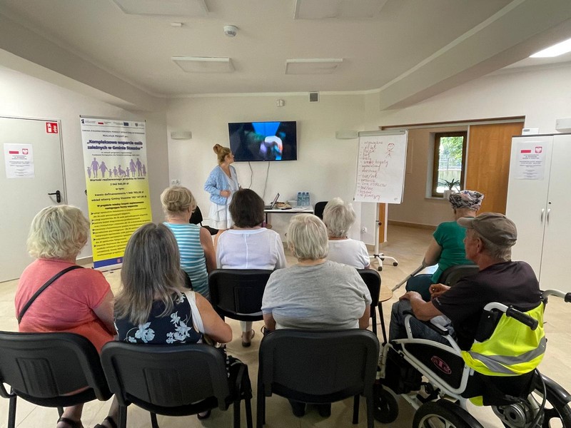 Grupa starszych osób siedzi na krzesłach oglądają film wyświetlany na telewizorze- Mężczyzna siedzi na wózku inwalidzkim
