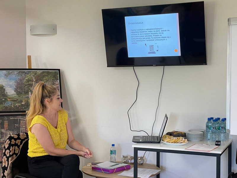 Pani w żółtej koszulce zwrócona w stronę laptopa który jest podpięty do telewizora na którym jest wyświetlany slajd