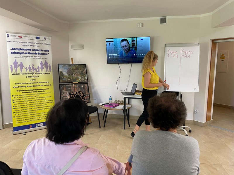 Pani w żółtej koszulce i czarnych jeansach stoi przy tablicy przed nia dwie kobiety siedzą odwrócone w jej kierunku