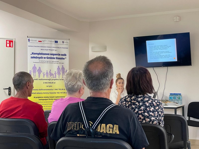 Grupa osób siedząca tyłem do obiektywu, ogladają slajd wyświetlany na telewizorze przed nimi młoda kobieta z zamyśloną miną