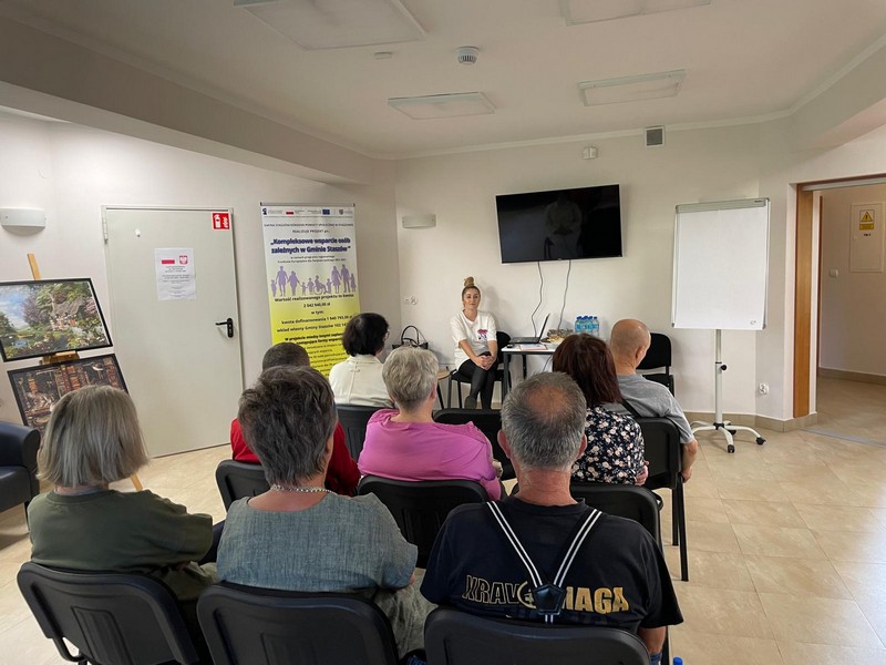 Grupa osób siedzi na krzesłach w dużym pomieszczeniu przed nimi młoda kobieta siedzi przy TV