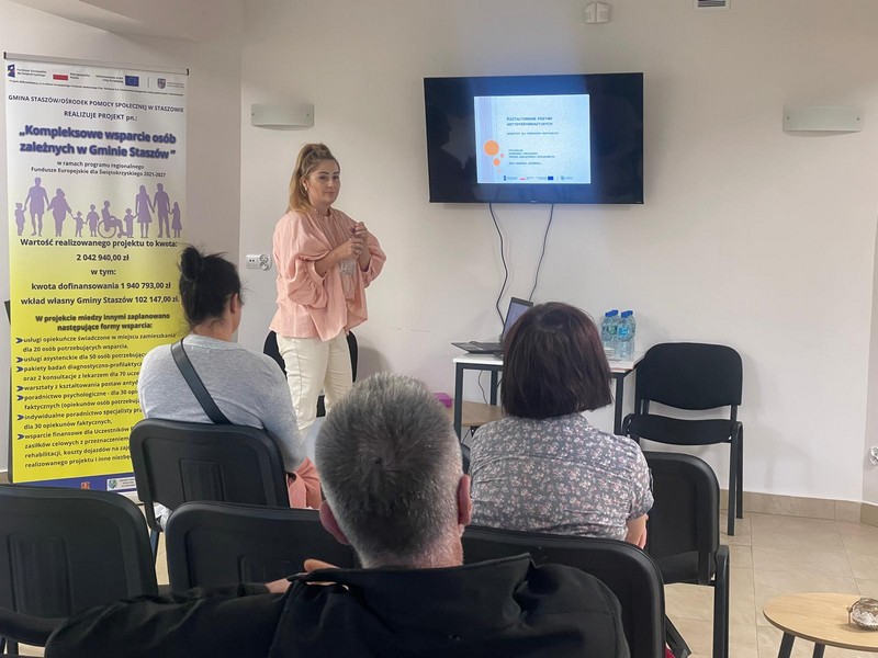 Kobieta w jasnoróżowej koszuli stoi przodem do grupki osób, które siedzą na krzesłach