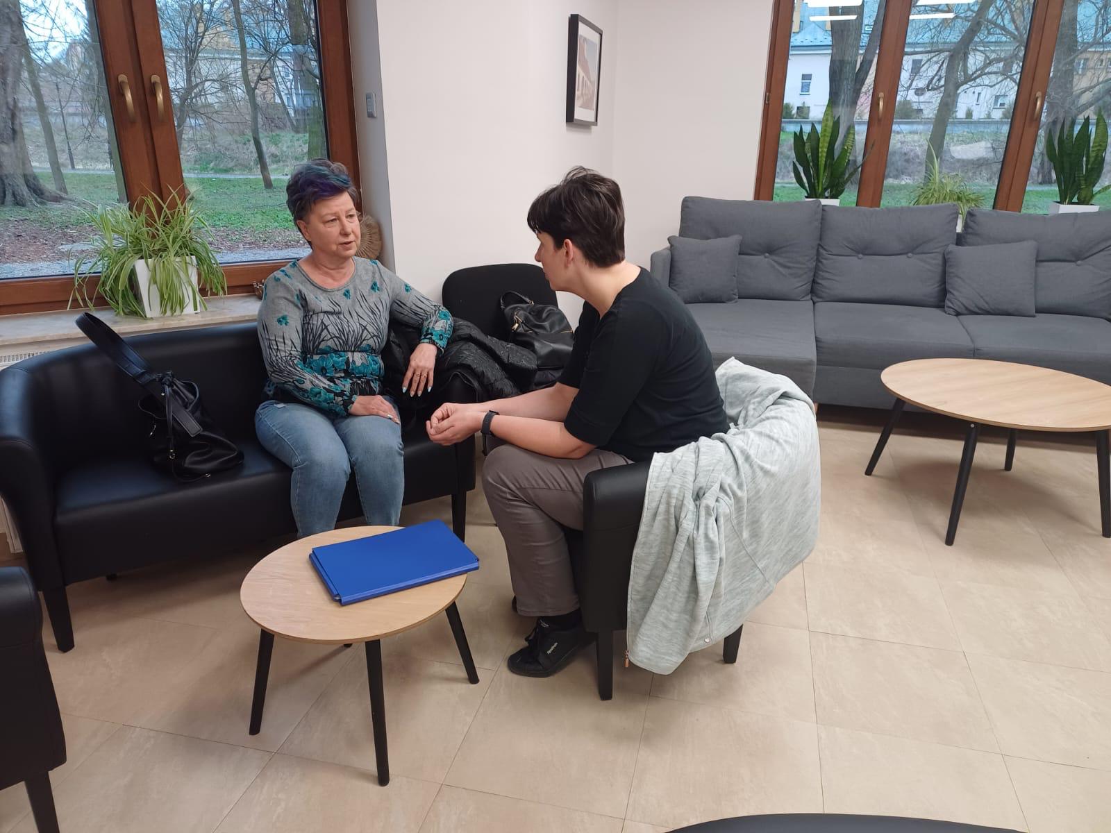 Dwie kobiety siedzą na wygodnych skórzanych fotelach i rozmawiają w tle okna przez które widać park.